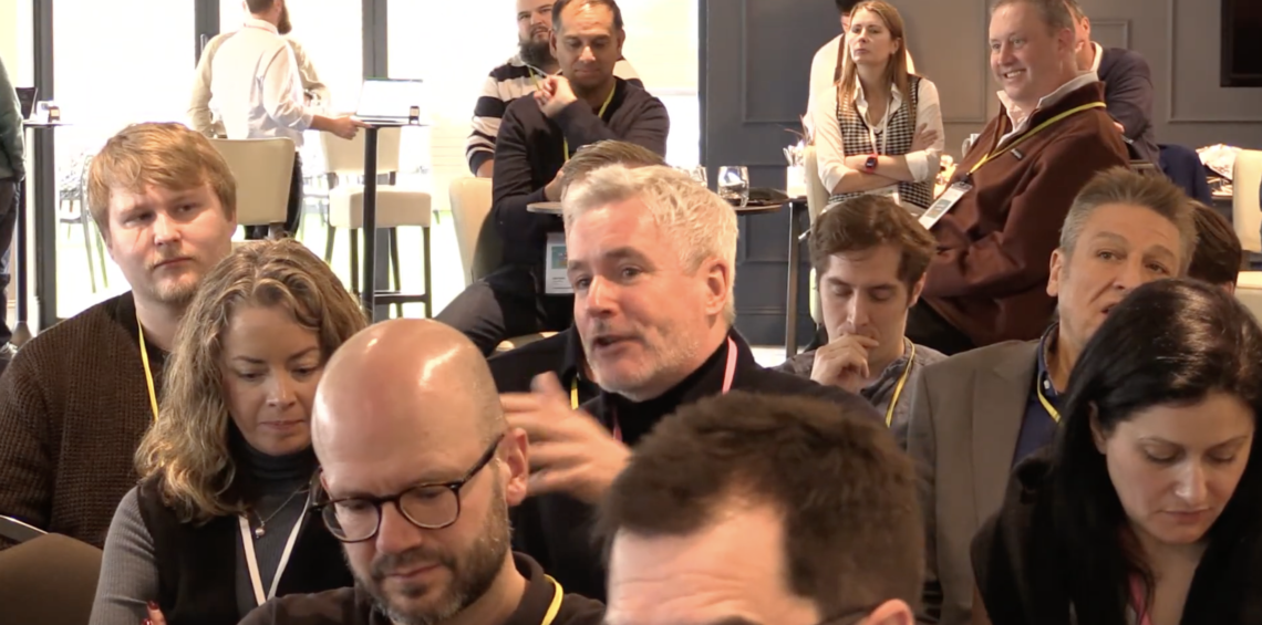 A man with a microphone in a seated audience asks a question while those around him listen.