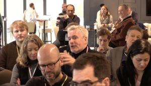 A man with a microphone in a seated audience asks a question while those around him listen.