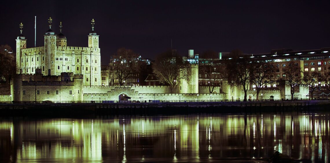 Photo of the Tower of London illuminated by floodlights against a dark nighttime sky and reflected in the River Thames
