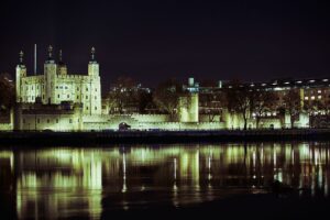 Photo of the Tower of London illuminated by floodlights against a dark nighttime sky and reflected in the River Thames